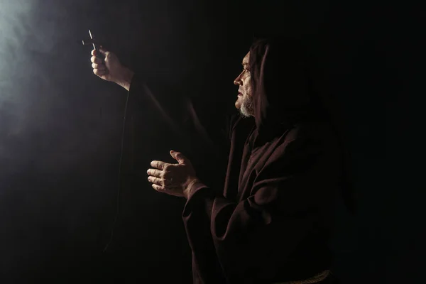 Monje en bata negra con capucha sosteniendo crucifijo santo en mano extendida sobre fondo oscuro - foto de stock