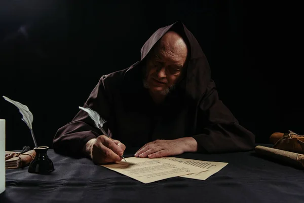 Medieval abbot writing chronicle on parchment near inkpot isolated on black — Stock Photo