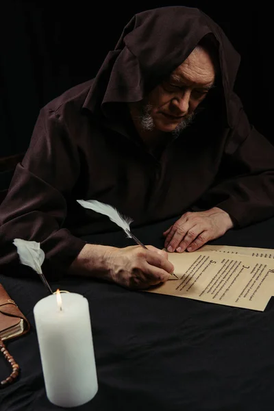 Monk in hooded robe writing on parchment near burning candle at night isolated on black — Stock Photo