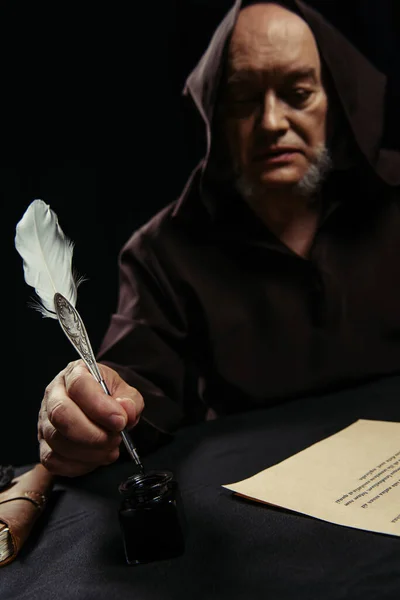 Blurred monk holding quill pen near ancient manuscript isolated on black — Stock Photo