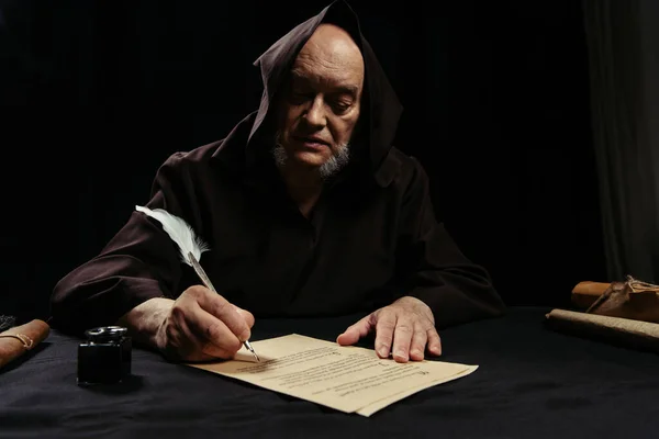 Priest in hooded robe writing chronicle on parchment isolated on black — Stock Photo
