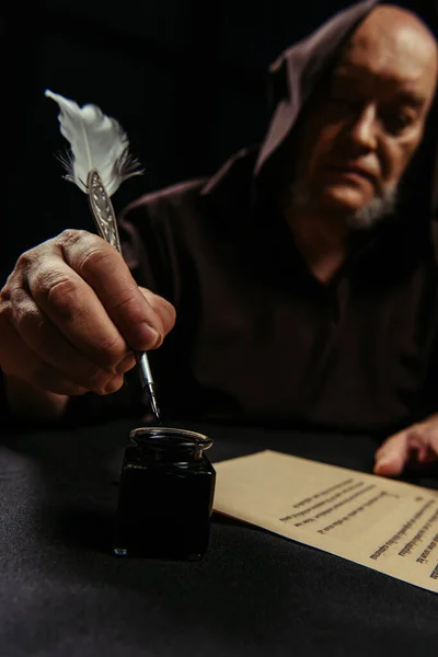 Blurred monk with quill pen near inkpot and manuscript isolated on black — Stock Photo