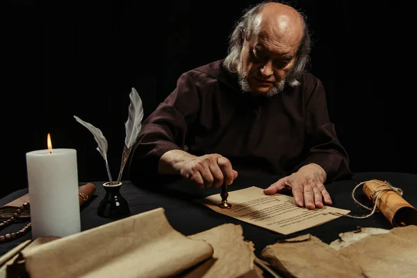 Senior abbot stamping manuscript with wax seal near parchments and burning candle isolated on black — Stock Photo