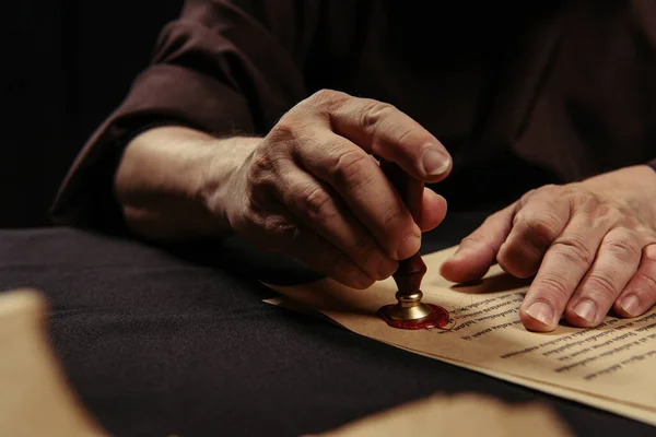 Cropped view of abbot stamping chronicle by wax seal on blurred foreground isolated on black — Stock Photo