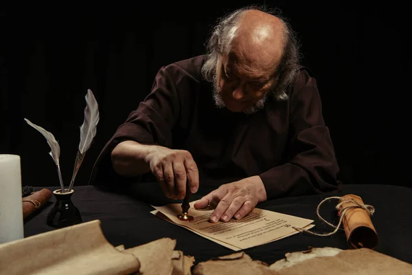 Senior monk stamping manuscript with wax seal near parchments isolated on black — Stock Photo