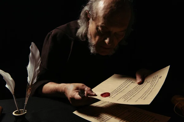 Senior priest holding wax seal and looking at stamped manuscript isolated on black — Stock Photo