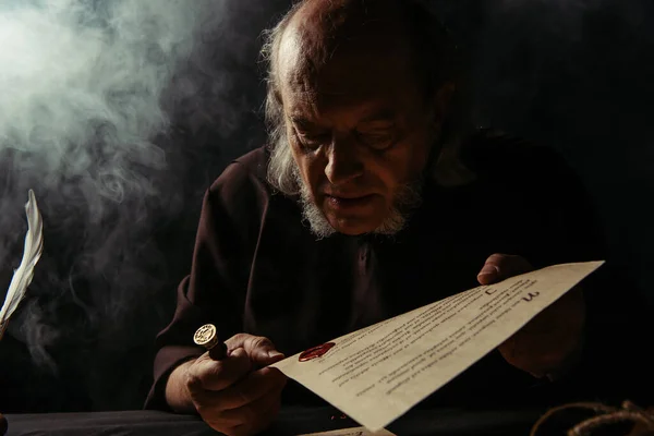 Senior priest looking at wax seal on manuscript on dark background with smoke — Stock Photo