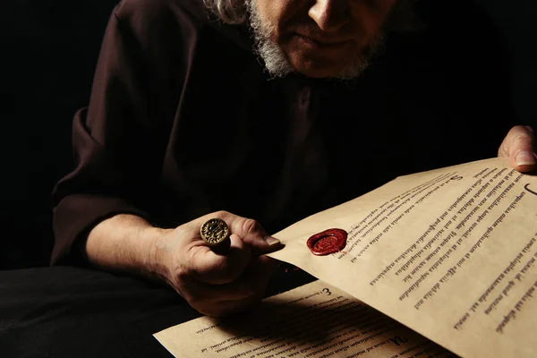 Vue recadrée du moine aîné tenant le parchemin avec le manuscrit et le sceau de cire isolé sur noir — Photo de stock