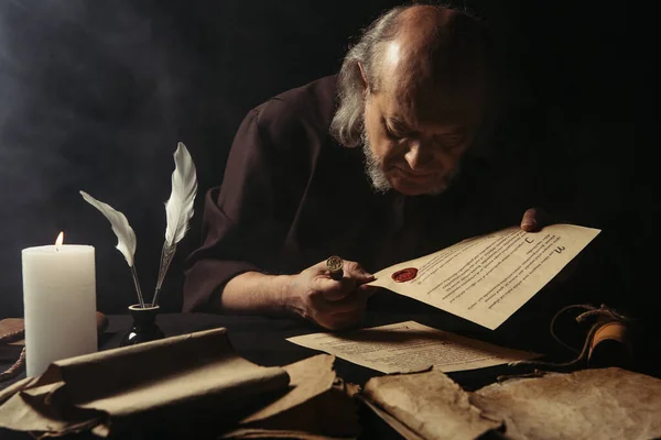 Senior monk looking at chronicle stamped with wax seal near parchments and burning candle on dark background — Stock Photo