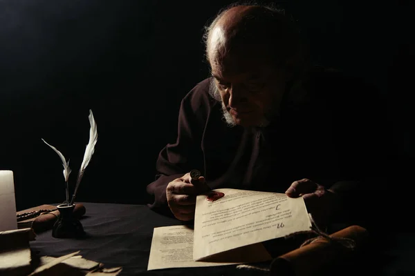 Monk holding wax seal and manuscripts in darkness isolated on black — Stock Photo