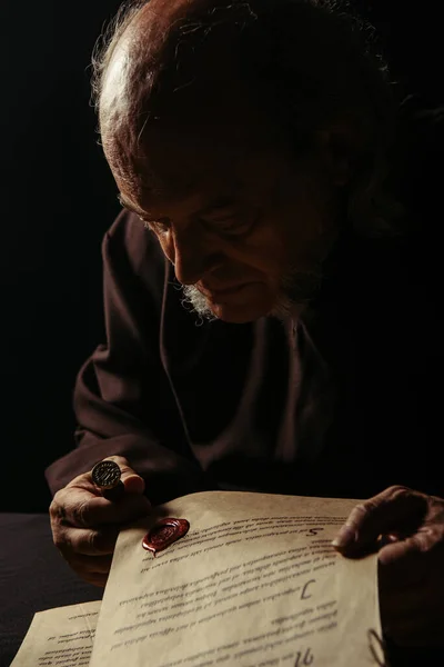 Середньовічний священик, дивлячись на воскову печатку на старовинному рукописі, ізольованому на чорному — стокове фото