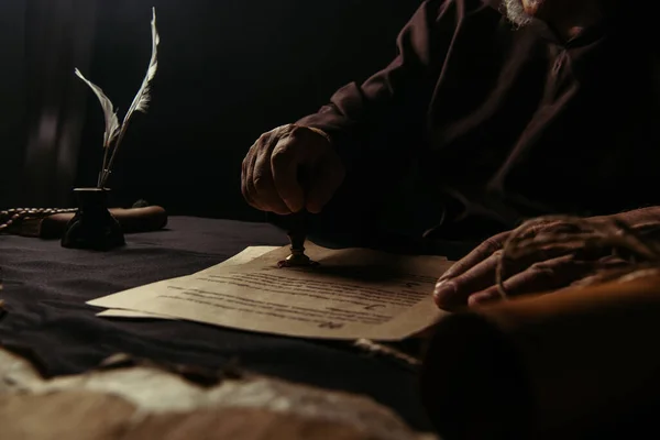 Ausgeschnittene Ansicht eines älteren Mönchs, der Wachssiegel auf Chronik in der Nähe von Tintenfass auf schwarzem Hintergrund stempelt — Stockfoto