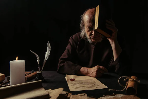 Senior priest obscuring face with bible near manuscripts stamped with wax seal  isolated on black — Stock Photo