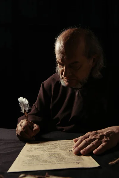 Mysterious monk writing chronicle with quill pen in darkness isolated on black — Stock Photo