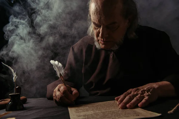 Monk in black cassock writing manuscript on dark background with smoke — Stock Photo