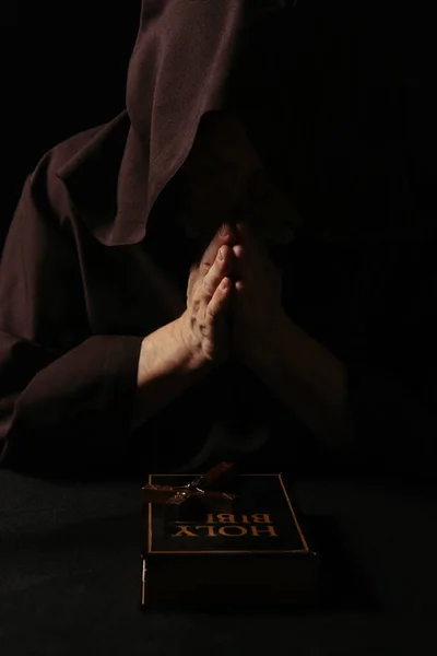 Anonymous monk praying near crucifix and holy bible isolated on black — Stock Photo