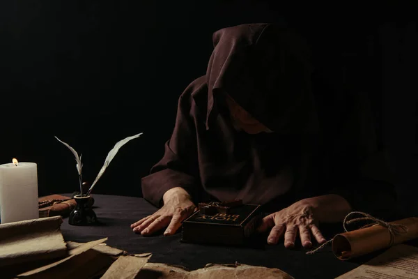 Senior abbot in hood sitting near bible and medieval manuscripts isolated on black — Stock Photo