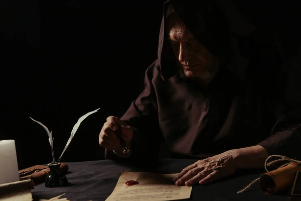 Senior abbot in hooded cassock stamping manuscript with wax seal isolated on black — Stock Photo