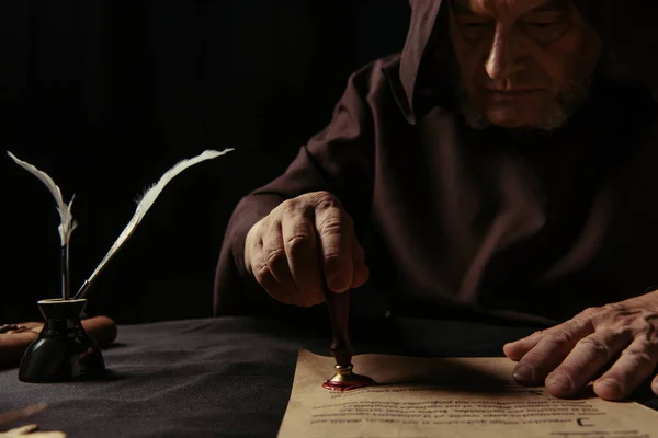 Середньовічний чернець в темному халаті з капюшоном, що штампує пергамент з восковою печаткою ізольовано на чорному — стокове фото