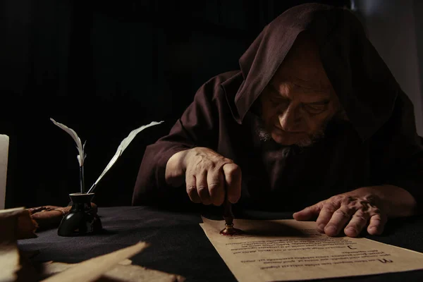 Mysterious monk in hood stamping manuscript with wax seal isolated on black — Stock Photo