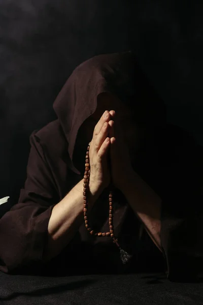 Anonymous monk in hooded robe praying with rosary beads isolated on black — Stock Photo