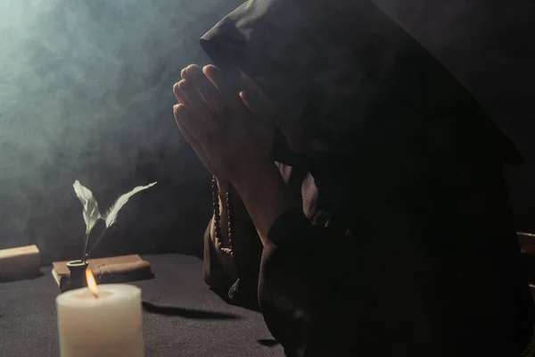 Medieval monk in hooded cassock praying with rosary beads near burning candle on black background with smoke — Stock Photo