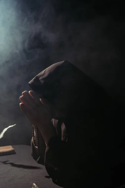 Monk with obscured face praying with rosary at night on black with smoke — Stock Photo