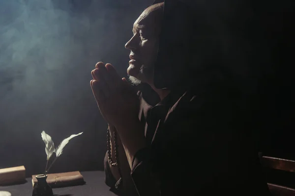 Medieval monk praying near inkpot and manuscript on black background with smoke — Stock Photo