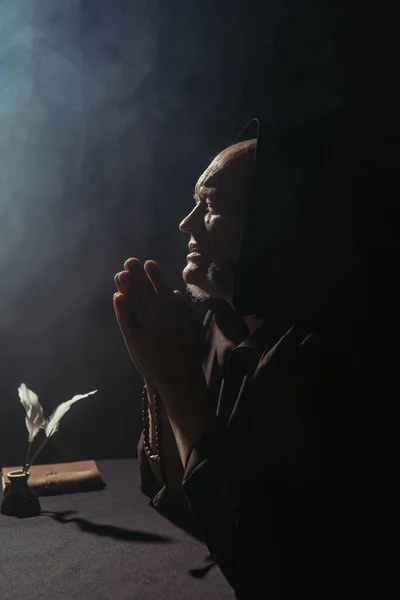 Senior priest with rosary beads praying near inkpot with quills on black with smoke — Stock Photo