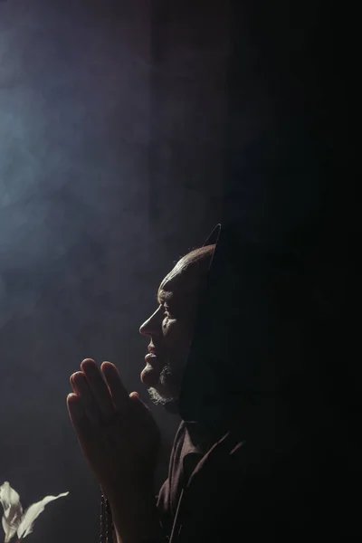 Side view of senior monk in hood praying at night on black background with copy space — Stock Photo