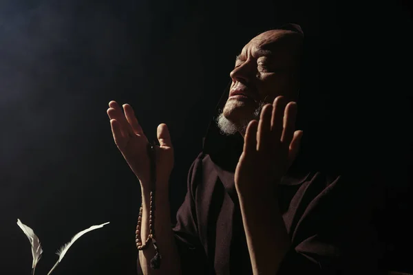Medieval priest praying with rosary and closed eyes isolated on black — Stock Photo