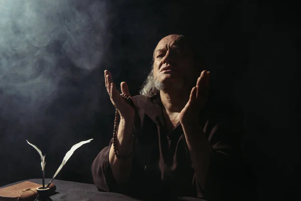 Medieval abbot with rosary praying near manuscript and inkpot with quills on black with smoke — Stock Photo