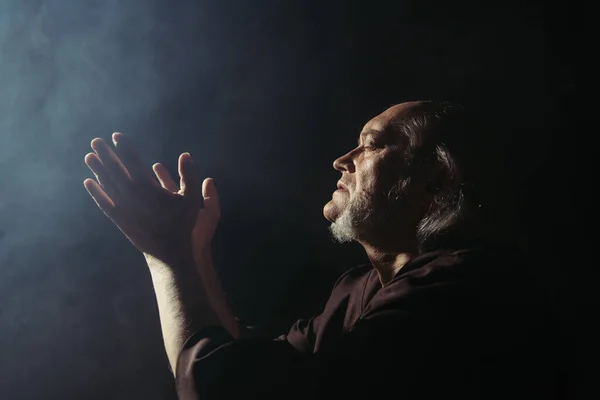 Side view of medieval priest praying on black background with smoke — Stock Photo