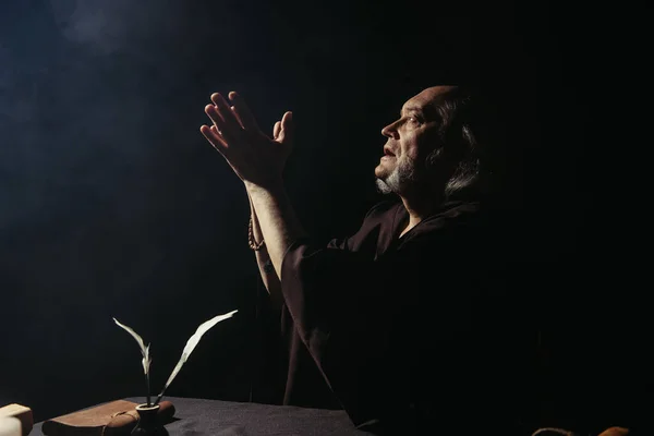 Side view of medieval monk praying near inkpot with quills on black background — Stock Photo