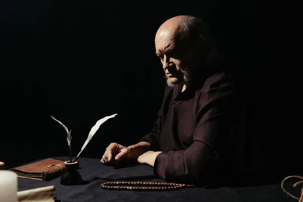 Thoughtful monk sitting near inkpot and rosary at night isolated on black — Stock Photo