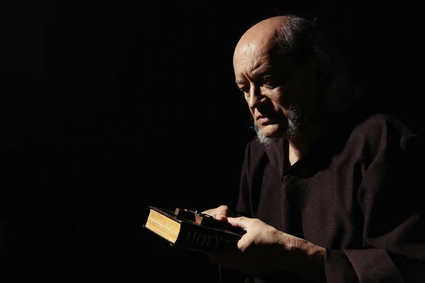 Monje mayor sosteniendo la Biblia y crucifijo santo aislado en negro - foto de stock