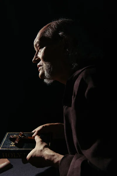 Vue latérale du prêtre aîné avec sainte bible et crucifix isolé sur noir — Photo de stock