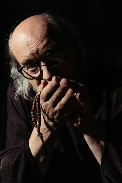 Senior monk in eyeglasses holding rosary and looking at camera isolated on black — Stock Photo