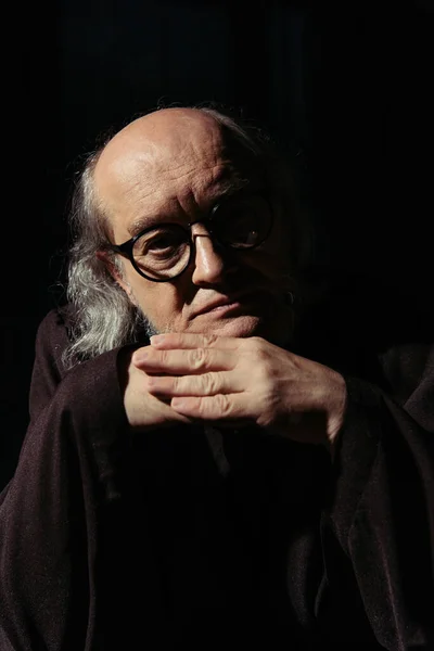 Religious philosopher in eyeglasses holding hands near chin and looking at camera isolated on black — Stock Photo