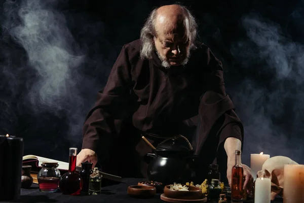 Medieval alchemist preparing potion near liquid and dried ingredients on black background with smoke — Stock Photo