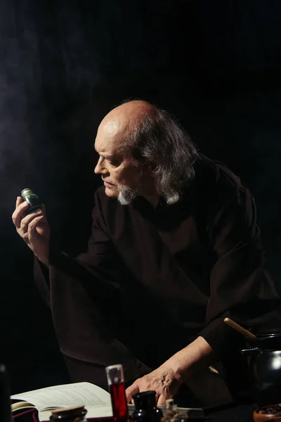 Senior alchemist holding vial with magic ingredient near flasks and magic cookbook isolated on black — Stock Photo