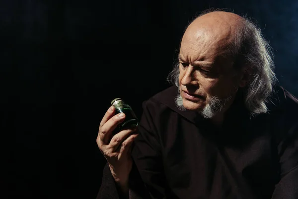 Alchimista pensieroso guardando vaso con ingrediente magico isolato su nero — Foto stock