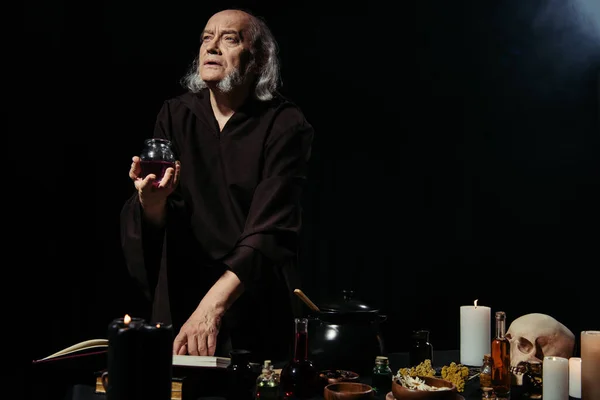 Thoughtful alchemist holding jar with liquid near magic cookbook on black background — Stock Photo