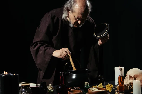 Alquimista en bata negra preparando poción con ingredientes herbarios y líquidos aislados en negro - foto de stock