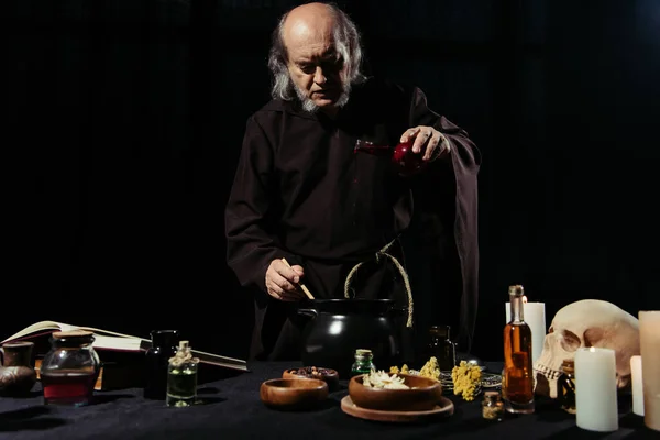 Senior alchemist adding ingredient into pot while preparing potion isolated on black — Stock Photo