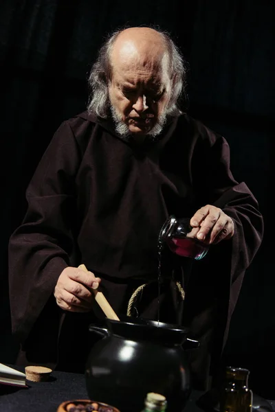 Alchemist in dark robe pouring liquid into pot while cooking at night isolated on black — Stock Photo