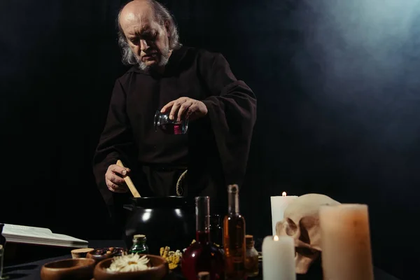 Mysteriöser Alchemist fügt Zutat in Topf beim Kochen in Dunkelheit auf schwarzem Hintergrund mit Rauch hinzu — Stockfoto