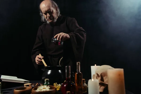 Mysterious alchemist adding ingredient into pot while cooking in dark on black background with smoke — Stock Photo