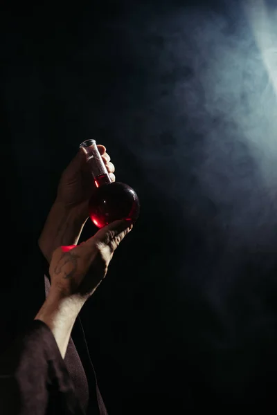 Partial view of magician holding flask with potion on black background with smoke — Stock Photo