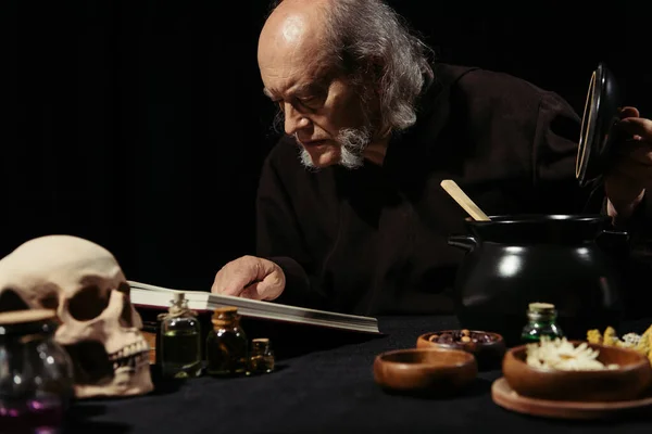 Asistente senior leyendo libro de cocina mágica cerca de olla e ingredientes aislados en negro - foto de stock